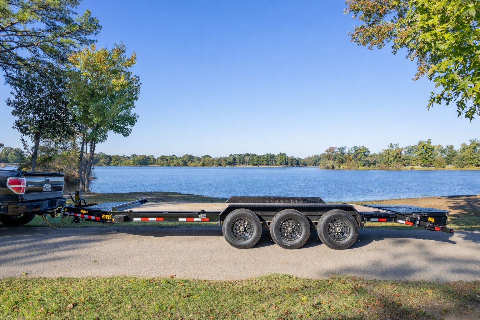 Black pickup truck towing a long, low flatbed trailer with three axles, parked on a paved road beside a calm lake, with trees and clear blue sky in the background.